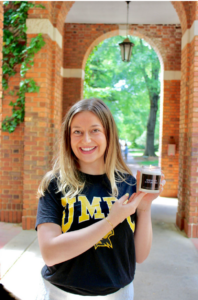 Mandi Lichtenstein holding a candle that says "Smells like a future PhD" and with a UMBC shirt